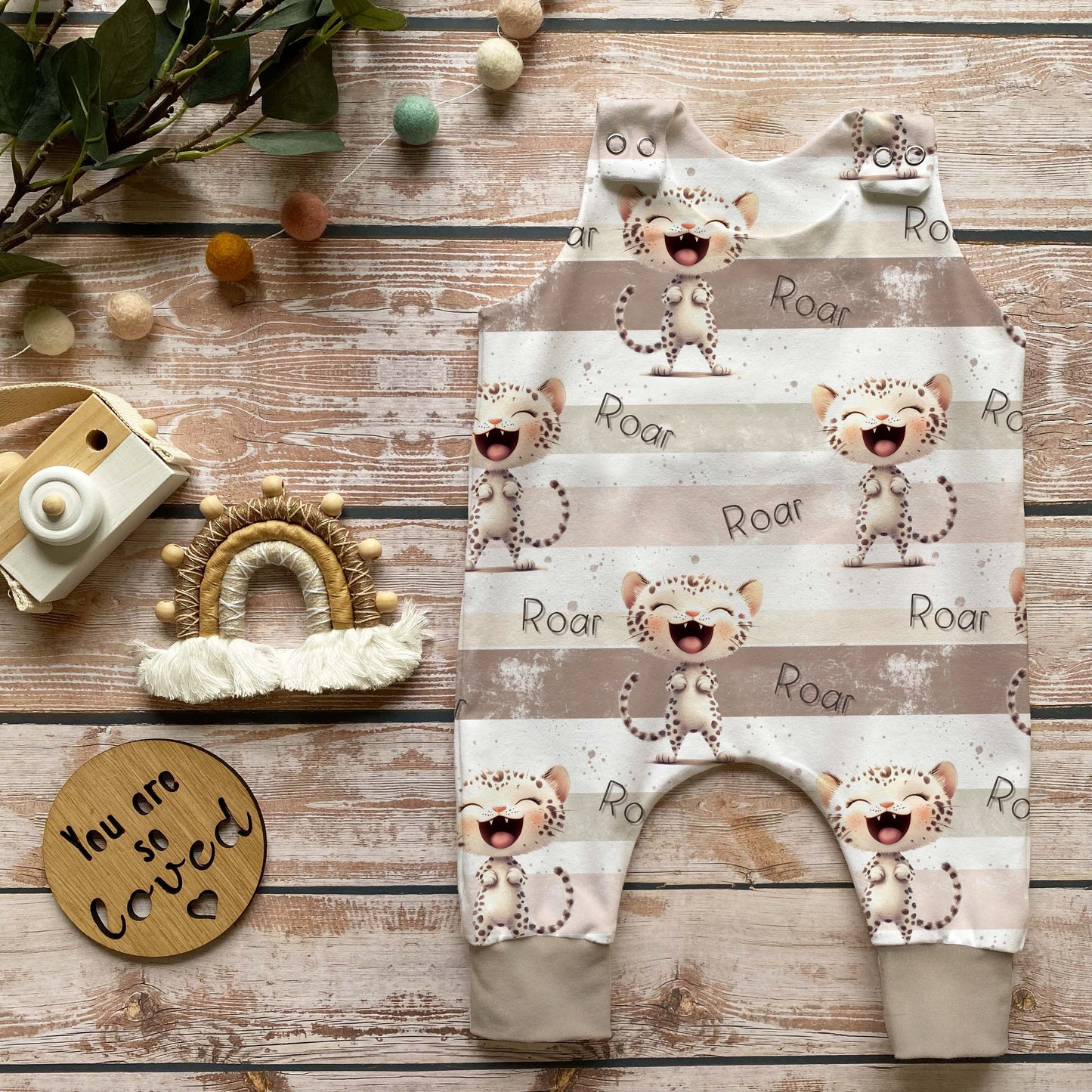 Handgenähter Baby-Strampler im Leo-Design in Beige mit „Roar“-Druck, weich und bequem aus French Terry, perfekt für Babys – liebevoll dekoriert mit Holzkamera, Makramee-Regenbogen und Naturdekoration.