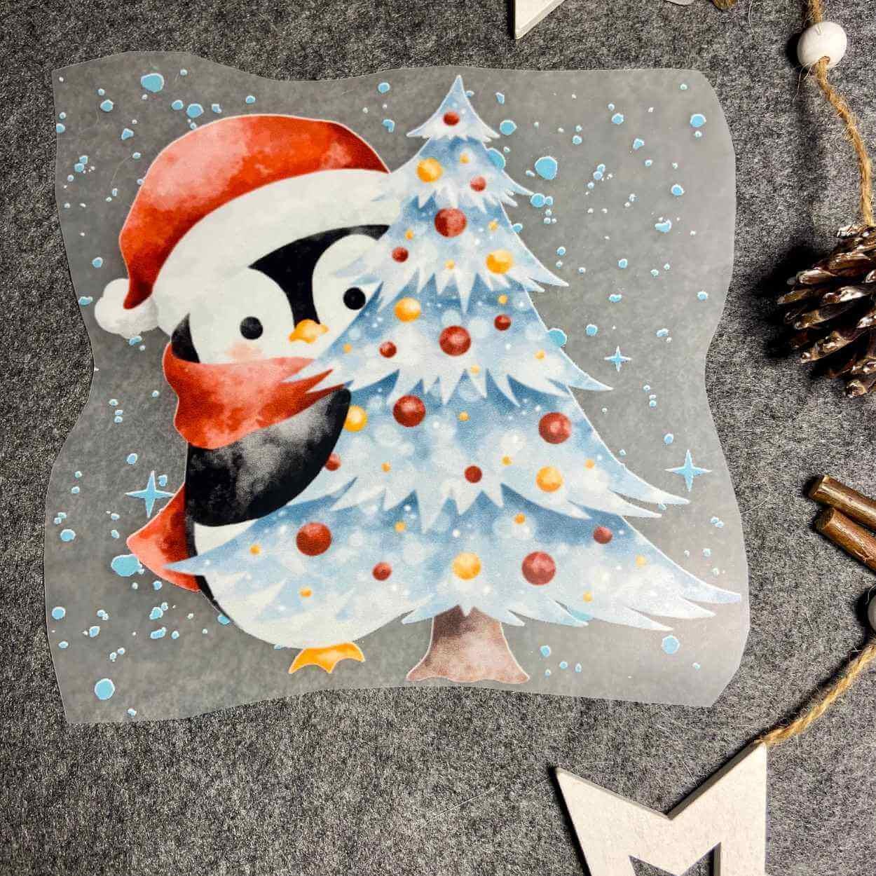 Bügelbild für Weihnachten, Pinguin im Schnee mit roter Weihnachtsmütze auf, einem roten Schal um und schaut hinter einem geschmücktem Tannenbaum hervor