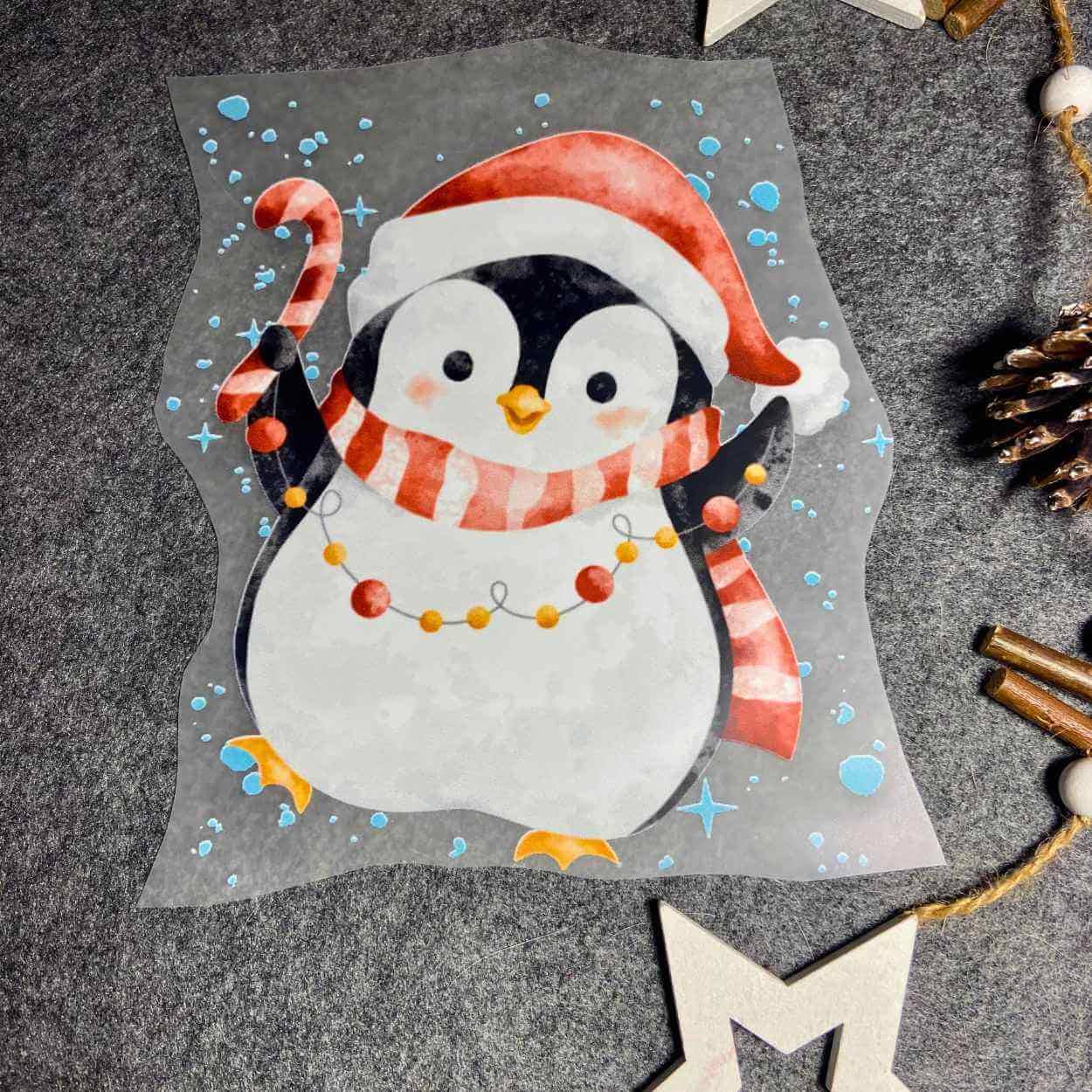 Weihnchtsbügelbild mit einem Pinguin mit Weihnachtsmütze auf und Schal um und einer Zuckerstange und Lichterkettte n der hand