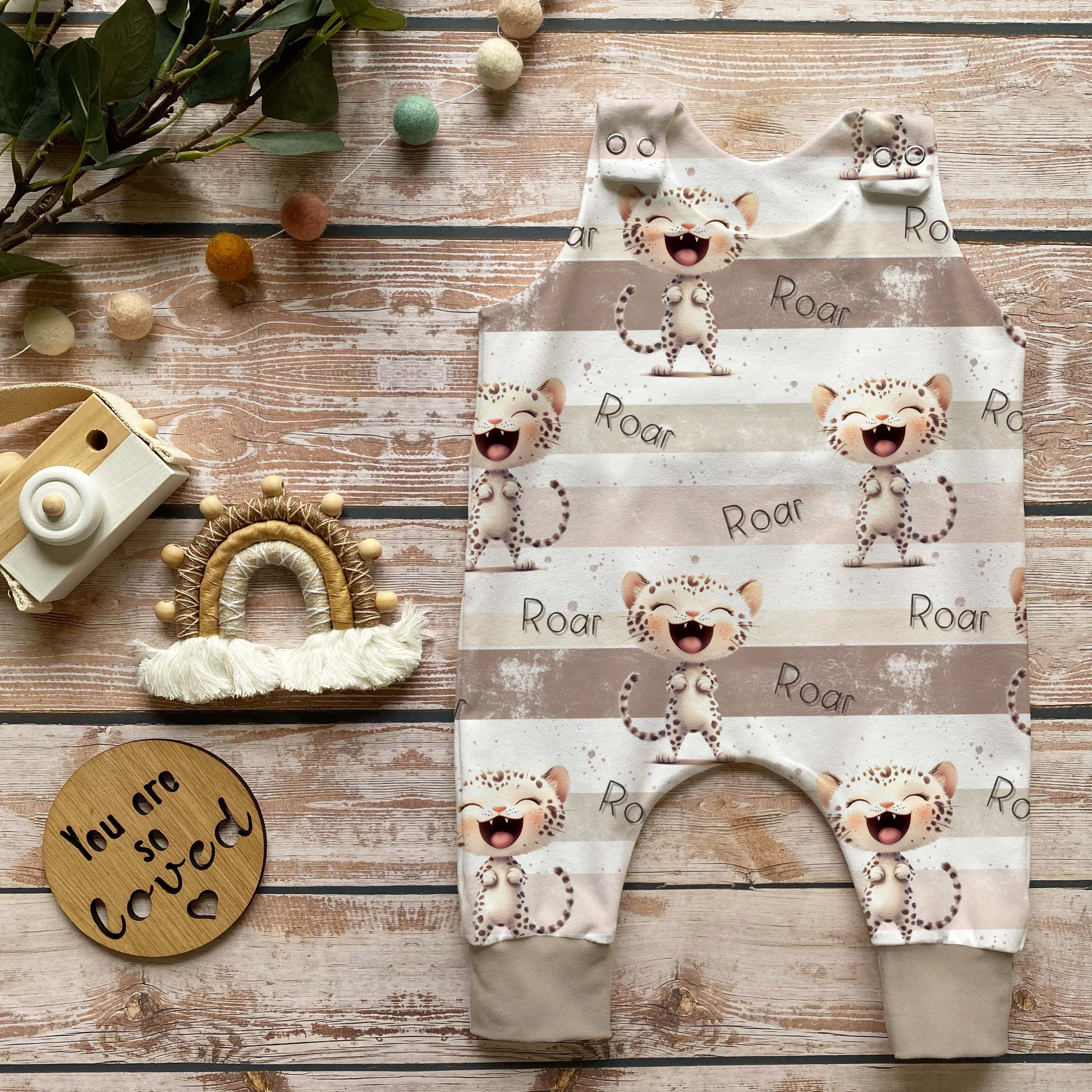 Handgenähter Baby-Strampler im Leo-Design in Beige mit „Roar“-Druck, weich und bequem aus French Terry, perfekt für Babys – liebevoll dekoriert mit Holzkamera, Makramee-Regenbogen und Naturdekoration.